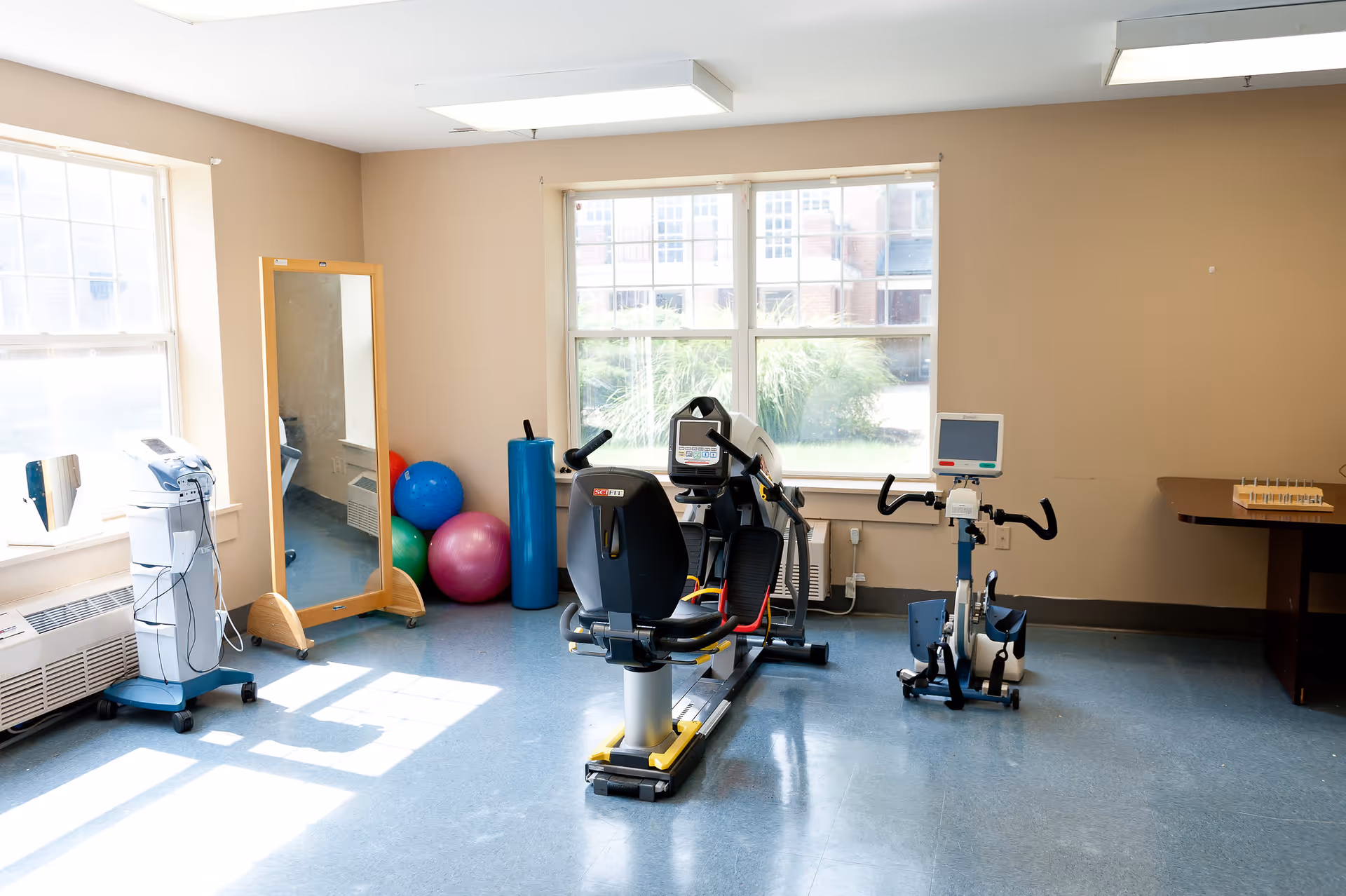 A bright room with exercise equipment including a recumbent stationary bike and a smaller exercise bike, colorful exercise balls, a tall mirror, and a medical device on a stand. The room has large windows letting in natural light and beige walls.