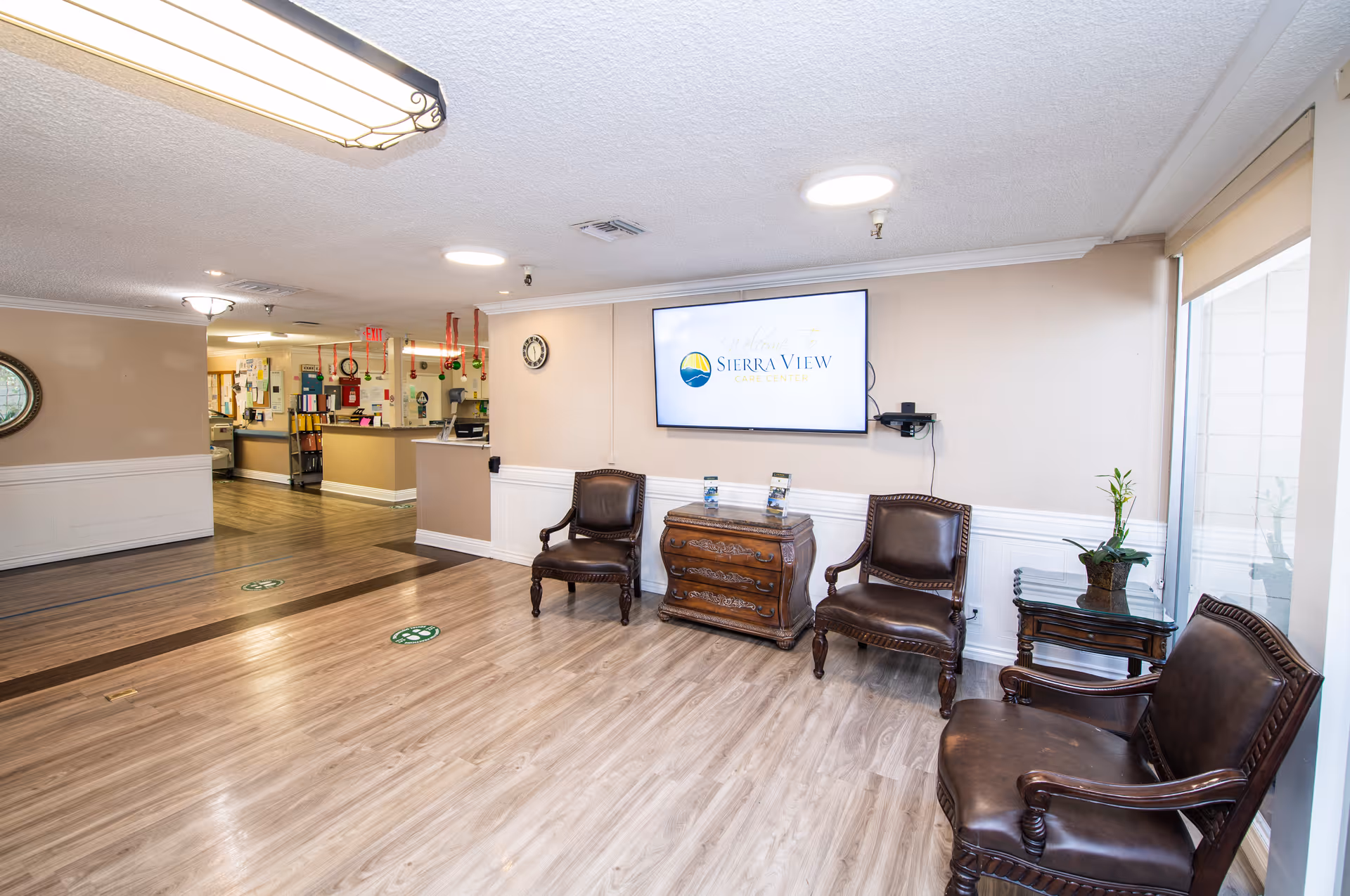 A clean and well-lit waiting area in Sierra View Care Center featuring three dark brown leather chairs, a wooden chest of drawers, a small side table with a plant, and a wall-mounted TV displaying the Sierra View Care Center logo. The floor has light wood laminate, and the reception desk is visible in the background.