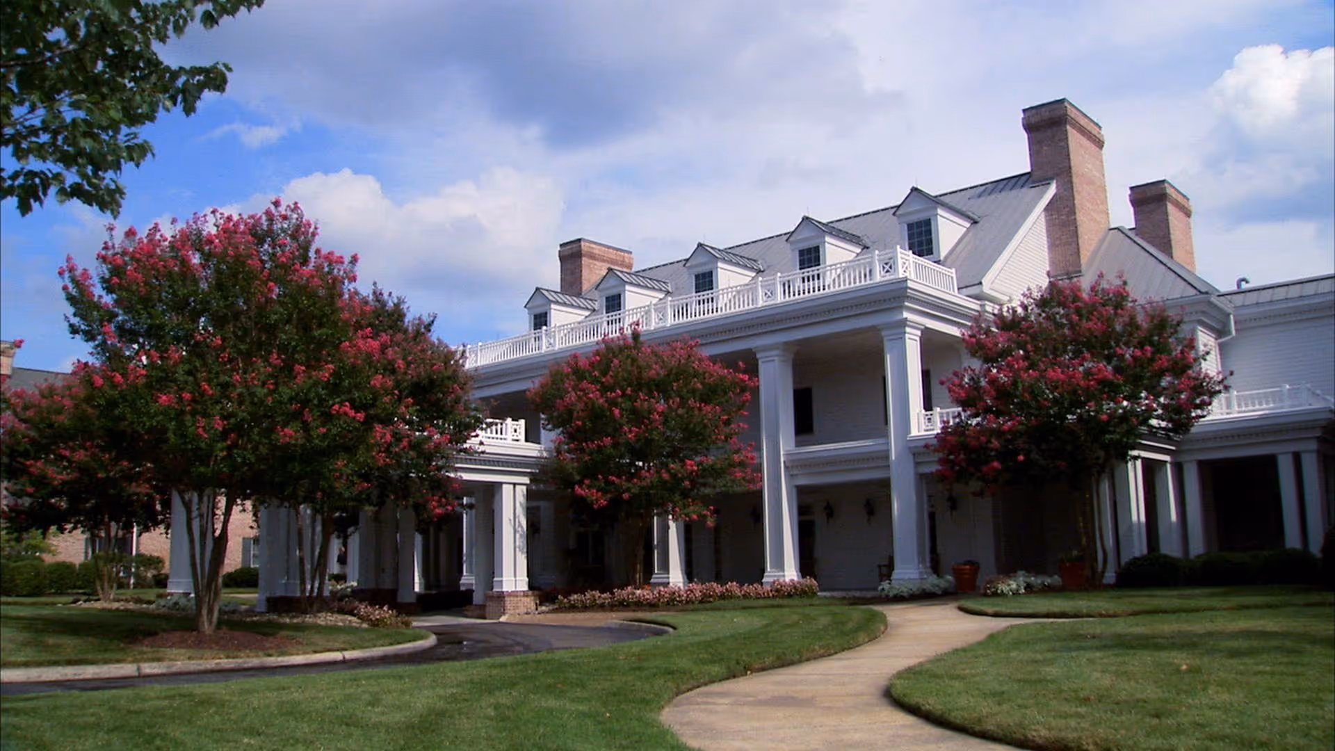 A grand white retirement facility with columns, dormer windows, and flowering trees along a curved path.