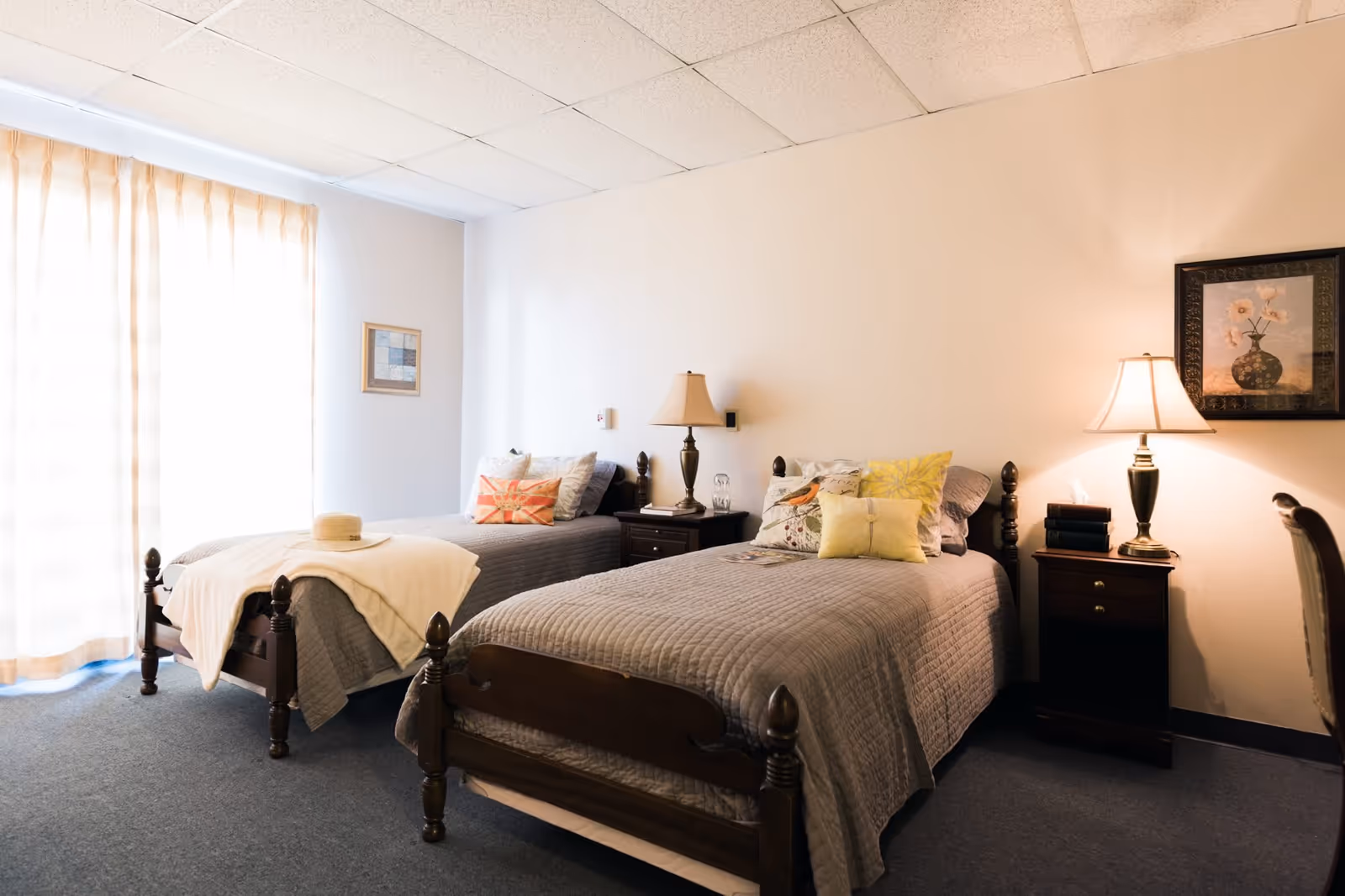 A cozy bedroom featuring two twin beds with gray quilts, decorative pillows, and bedside tables with lamps. Natural light filters through sheer curtains, illuminating the room's warm tones.