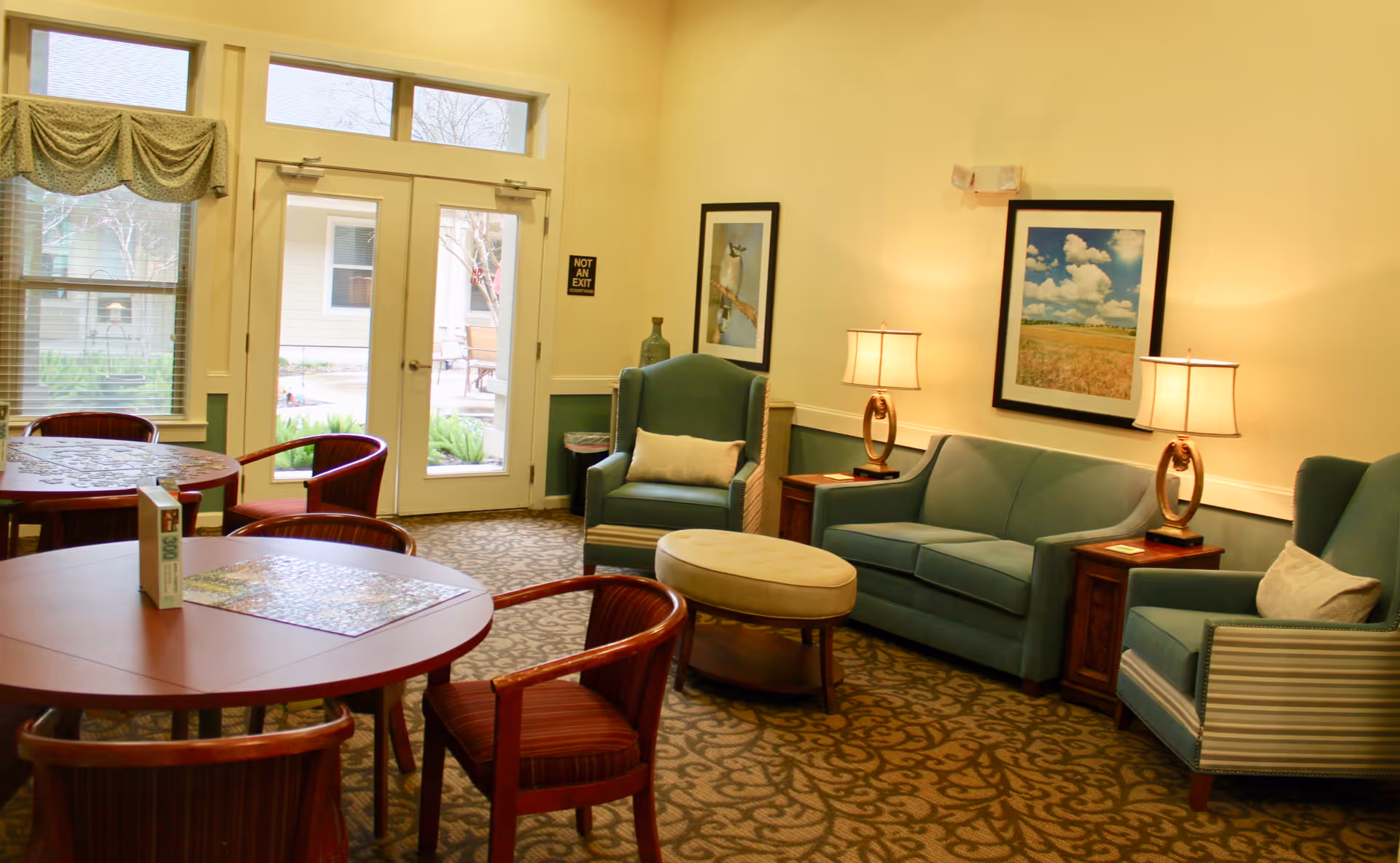 A cozy common area in an assisted living facility featuring two blue armchairs, a blue sofa, two side tables with lamps, framed artwork on the wall, and two round wooden tables with chairs. There are glass doors and windows letting in natural light, with a view of an outdoor patio area.