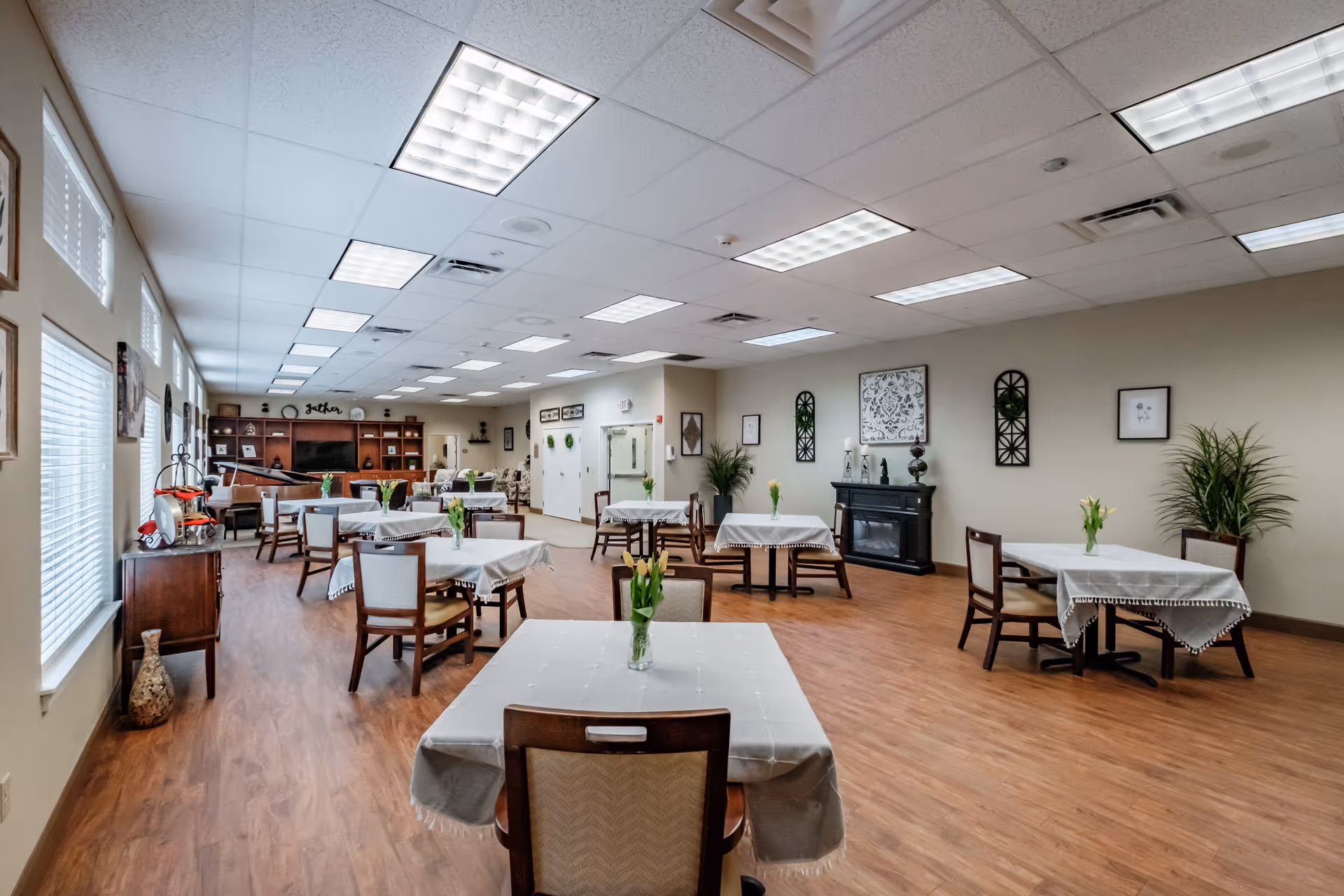 A spacious dining room with several tables covered with white tablecloths, each adorned with a small vase of yellow flowers. Wooden chairs surround the tables. The room has wood flooring, beige walls decorated with framed art and plants, and a ceiling with fluorescent lighting. At the far end, there is a large wooden shelving unit with a TV and decorative items.
