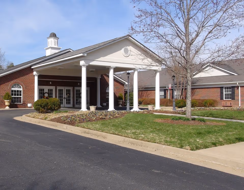 Exterior view of a senior living facility with a brick building, white columns supporting a covered entrance, a small landscaped garden area, and a leafless tree on a clear day.