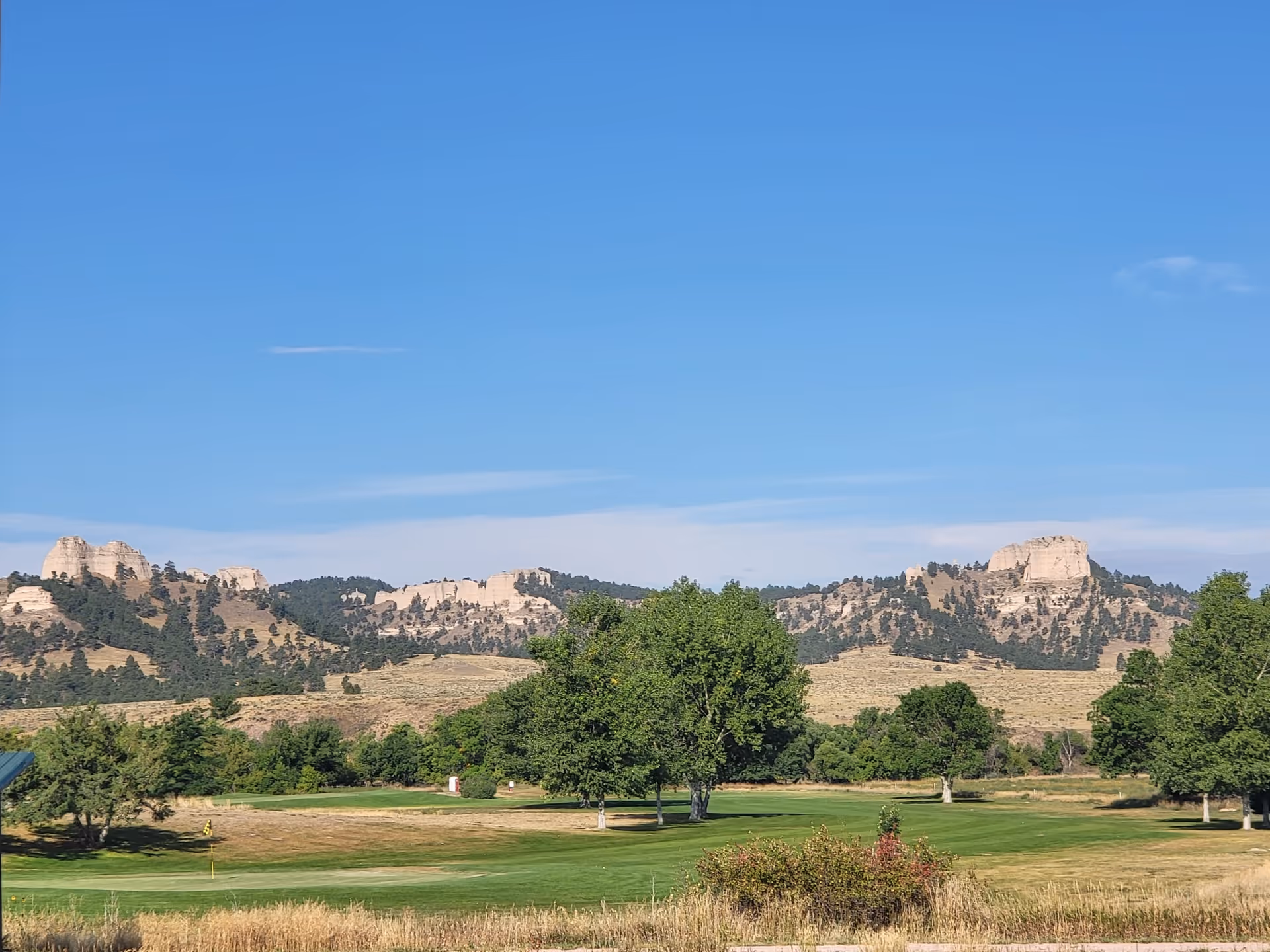 A scenic outdoor view featuring a green grassy area with scattered trees in the foreground and rocky hills with sparse vegetation in the background under a clear blue sky.