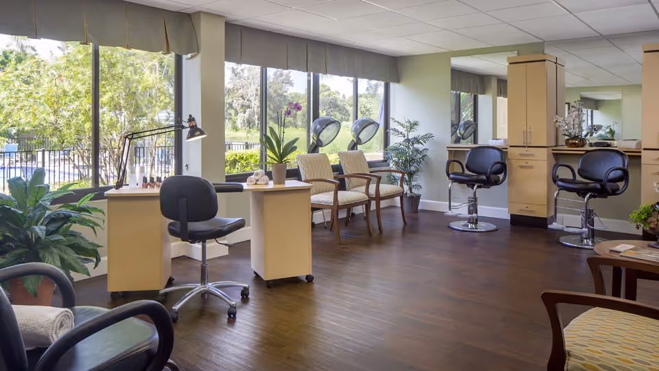 A bright and spacious salon area with large windows showing greenery outside. The room features salon chairs, hair drying stations, a manicure table with a lamp, potted plants, and comfortable waiting chairs. The floor is wooden, and the walls are painted light green.
