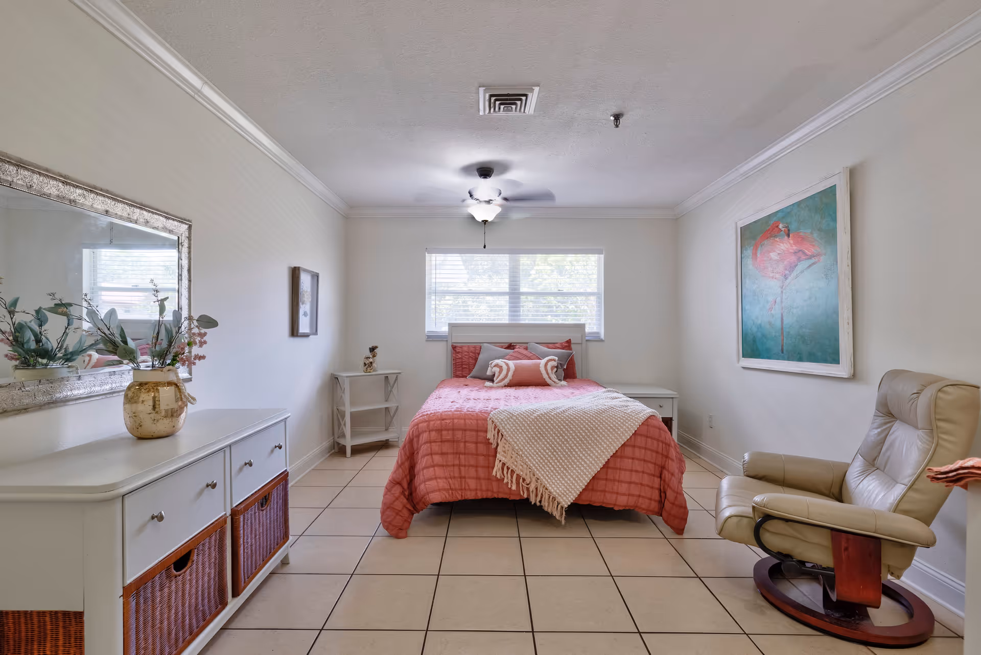 A bright bedroom with a bed covered in a coral quilt and decorative pillows, a beige recliner chair, a white dresser with woven baskets, a large mirror above the dresser, a small white side table, and a large painting of a pink flamingo on the wall. The room has tiled floors and a ceiling fan with a light fixture.