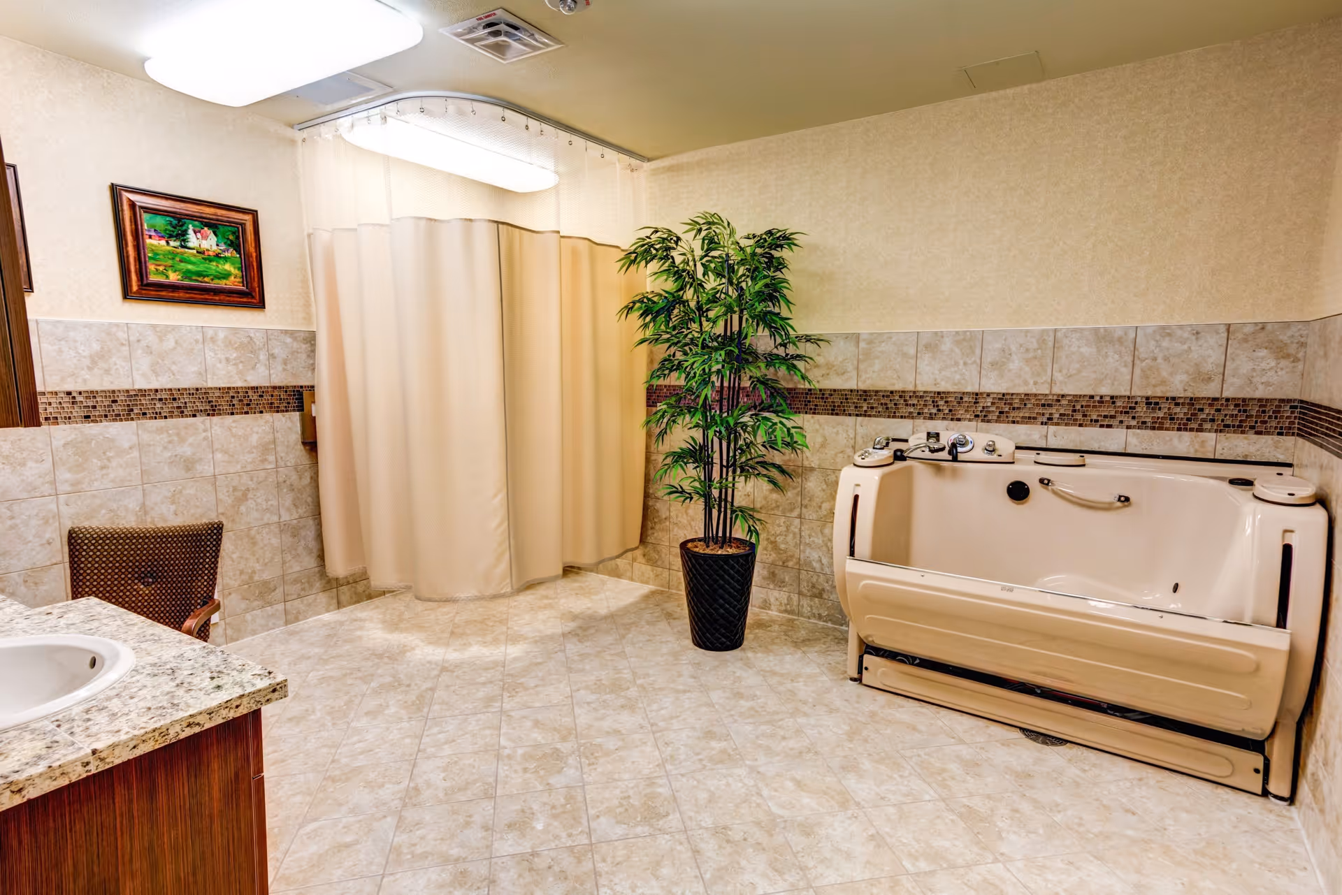 A spacious bathroom with beige tiled floors and walls, featuring a walk-in bathtub with safety handles, a sink with a granite countertop, a brown chair, a potted green plant, and a framed painting on the wall. A beige shower curtain hangs in the corner.