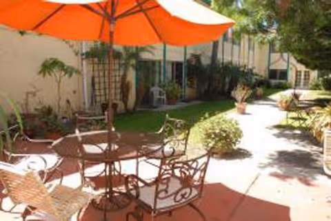 Outdoor patio area with a round metal table and several metal chairs under a large orange umbrella. The patio is adjacent to a building with windows and surrounded by green plants and trees.