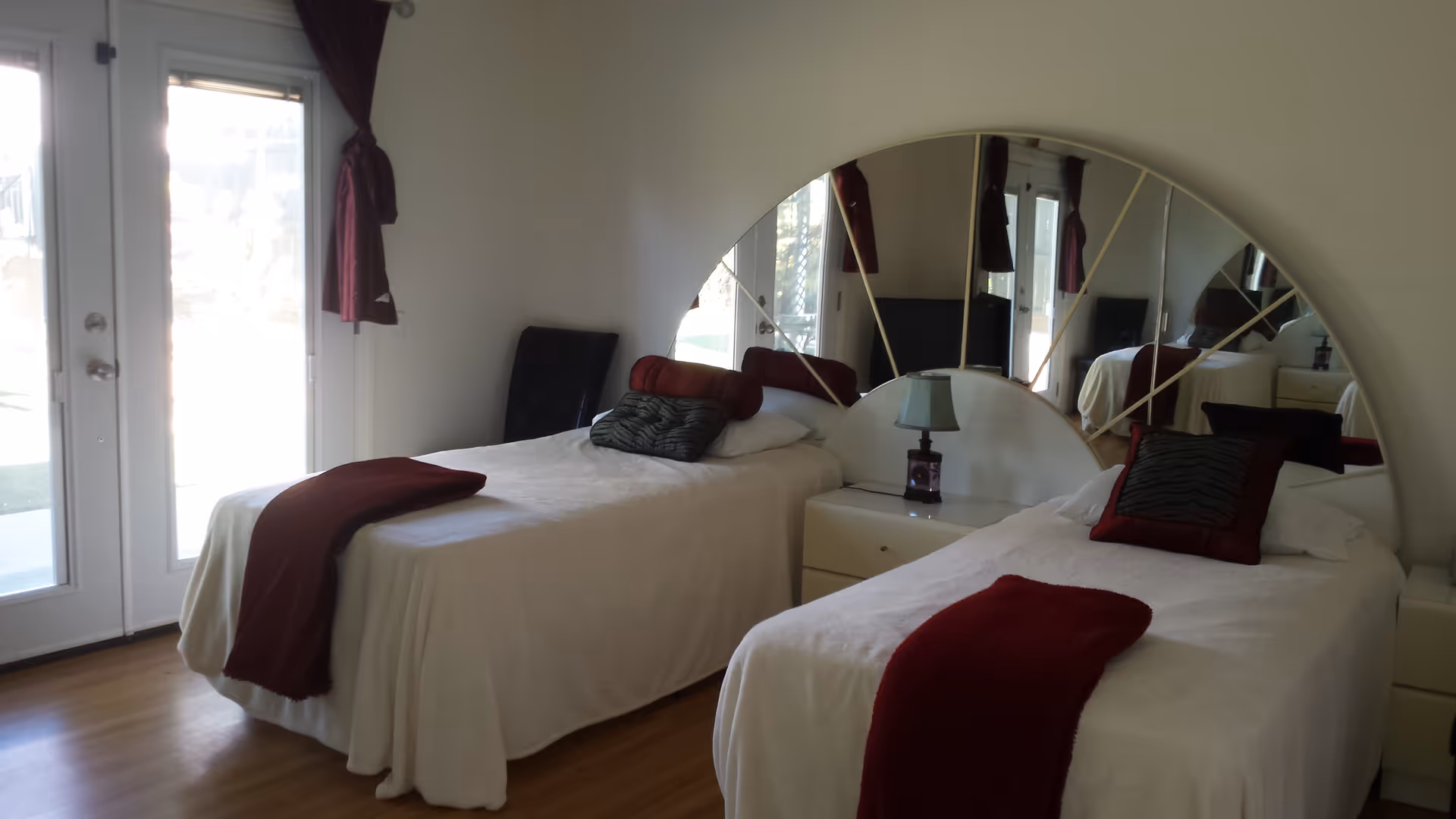 A bedroom with two single beds covered in white bedspreads and red throws. Each bed has decorative pillows. Between the beds is a white nightstand with a small lamp. Behind the nightstand is a large semi-circular mirror on the wall. To the left, there are glass doors with maroon curtains tied back, letting in natural light. The floor is wooden.
