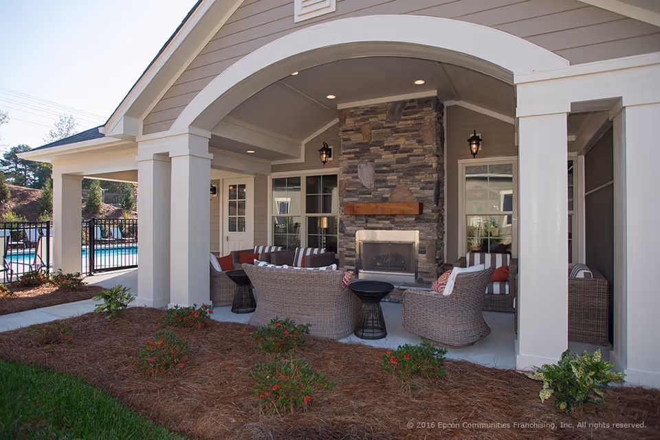 Covered outdoor seating area with wicker chairs and sofas arranged around a stone fireplace under a vaulted ceiling. The area is surrounded by white pillars and overlooks a pool area with a black metal fence. There are small plants and mulch beds in the foreground.