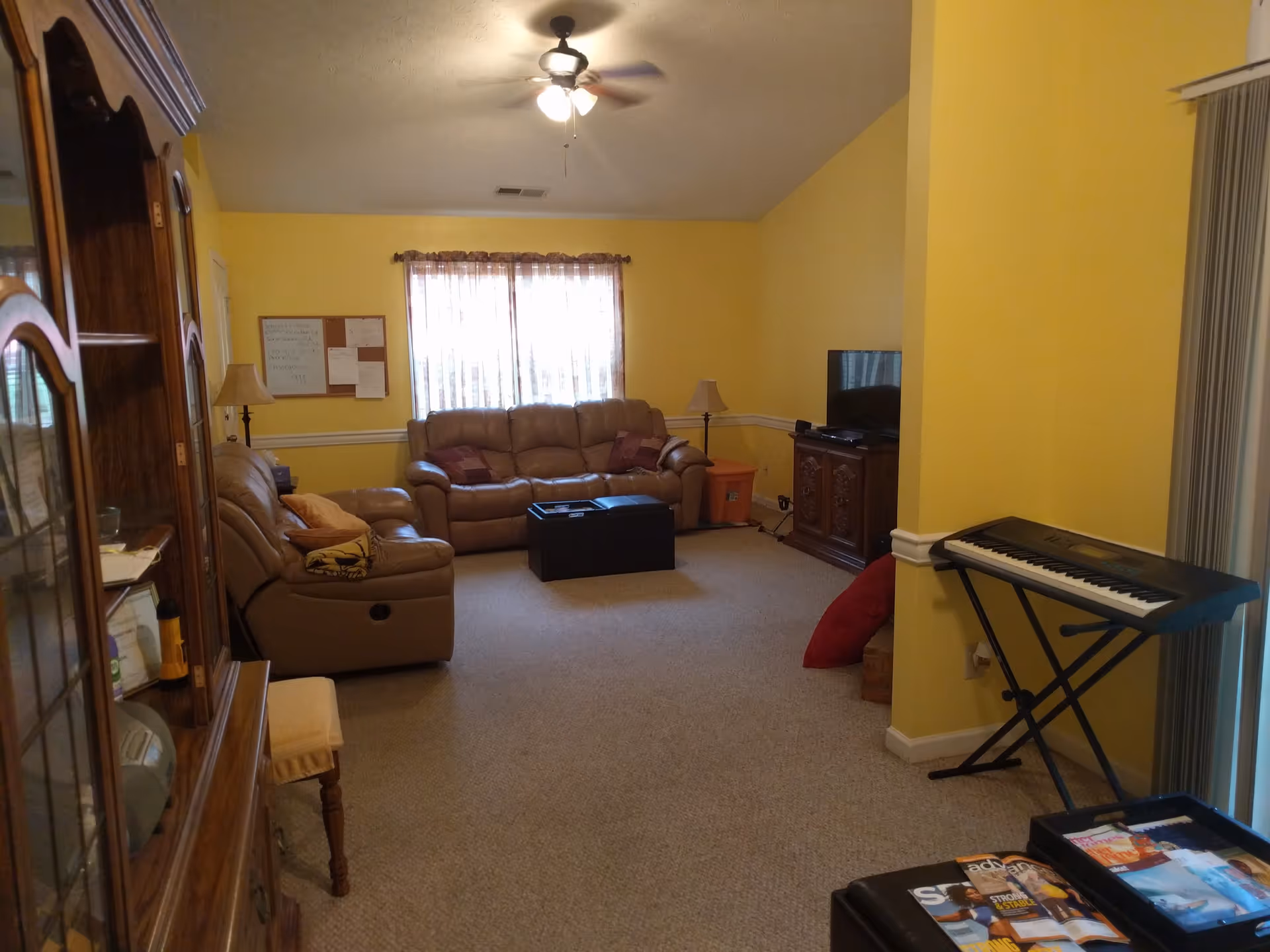 A cozy living room with yellow walls and beige carpet. The room features a brown leather sofa and loveseat, a wooden cabinet with a TV on top, a keyboard on a stand, a ceiling fan with lights, and a window with sheer curtains. There are also a couple of floor lamps, a small table with magazines, and a bulletin board on the wall.