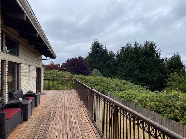 A wooden deck attached to a building with outdoor seating including chairs and small tables. The deck overlooks a lush green area with trees and bushes under a cloudy sky.