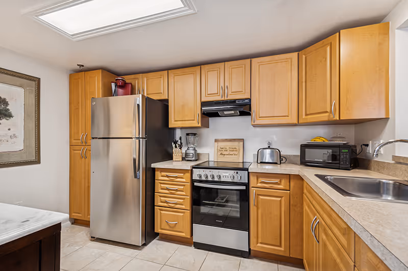A kitchen with light wood cabinets, a stainless steel refrigerator, an electric stove with oven, a blender, a toaster, a microwave, and a sink. There is a framed picture on the wall and a small sign on the countertop that reads 'Family, friends, love and laughter, always makes a house a home.'