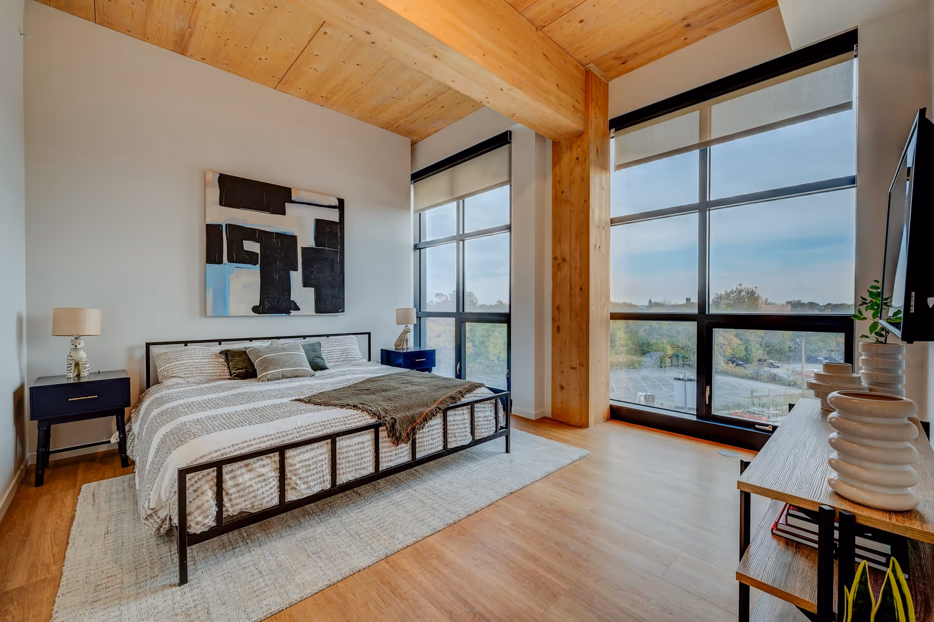 A modern bedroom with a large bed featuring a metal frame and striped bedding. There are two dark blue nightstands with lamps shaped like white tigers on either side of the bed. A large abstract black and white painting hangs above the bed. The room has wooden floors, a light-colored rug, and large floor-to-ceiling windows with roller shades, offering a view of trees and a parking lot outside. A wooden beam and ceiling add warmth to the space. A wooden console table with decorative white vases and a mounted flat-screen TV are visible on the right side.