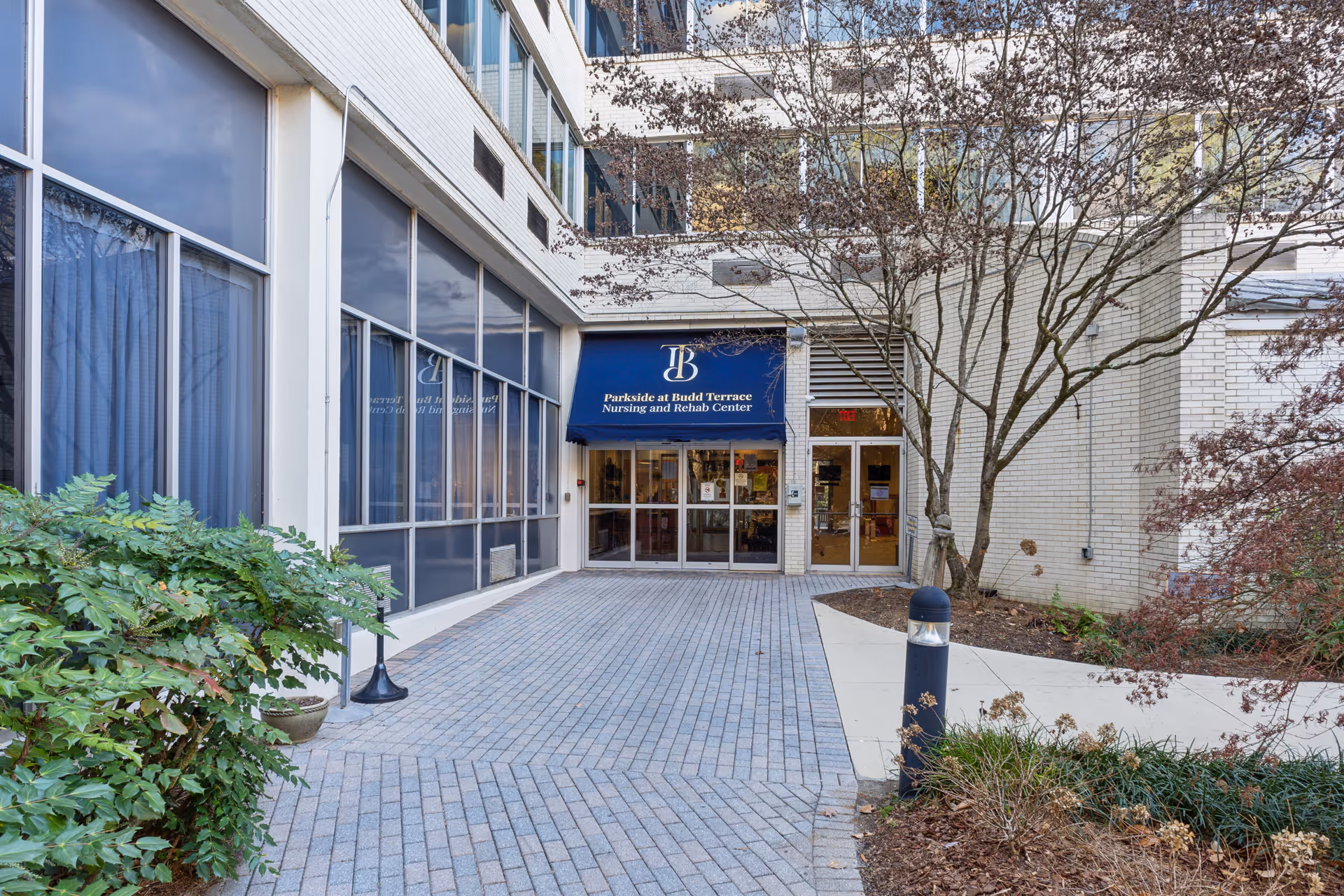 Entrance to Parkside at Budd Terrace Nursing and Rehab Center with a blue awning displaying the facility name and logo, surrounded by brick walls, large windows, trees, and landscaping along a paved walkway.