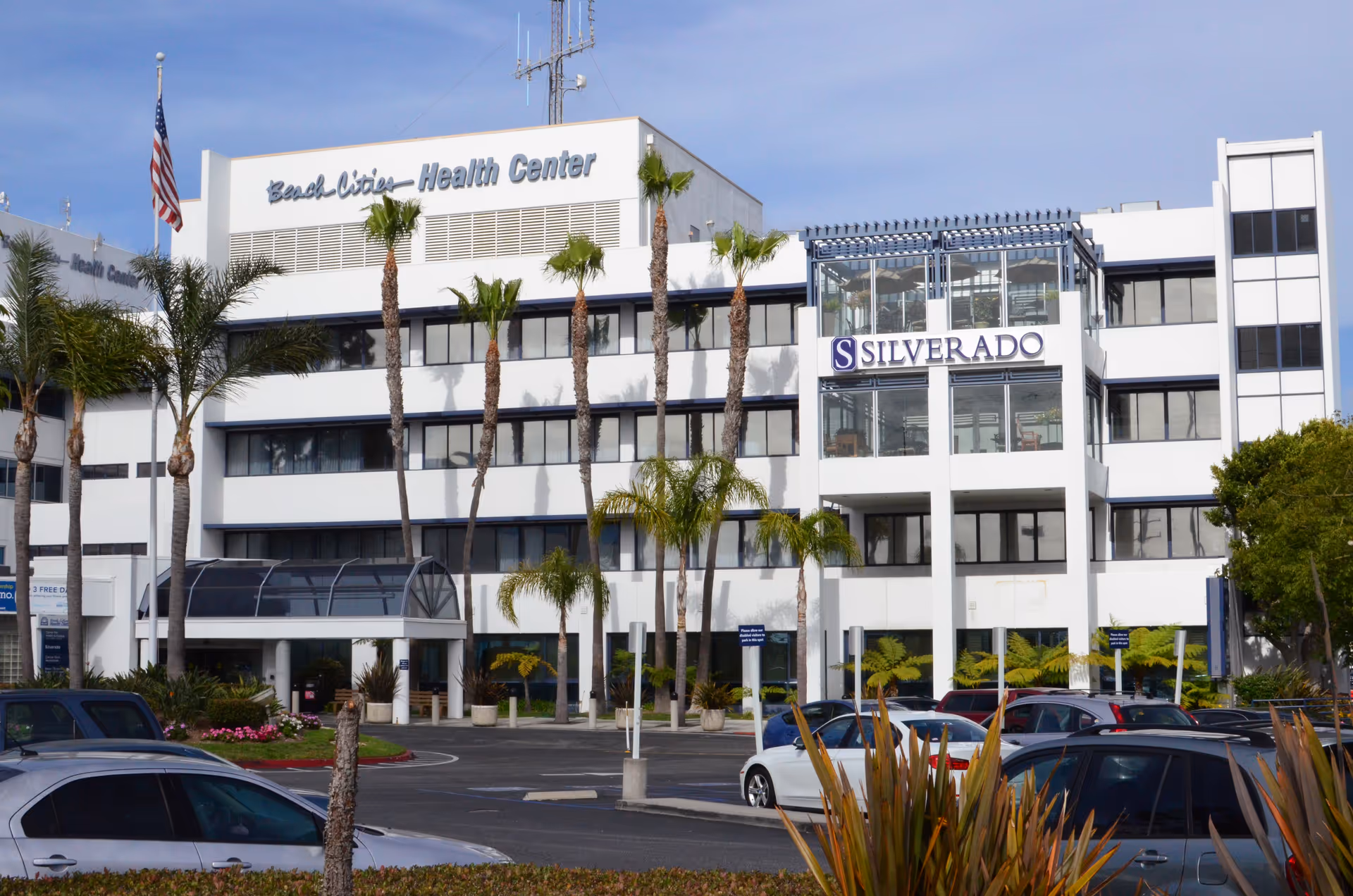 Exterior view of a multi-story white building with large windows and palm trees in front. The building has signs that read 'Beach Cities Health Center' and 'Silverado'. Several cars are parked in the parking lot in front of the building under a clear sky.