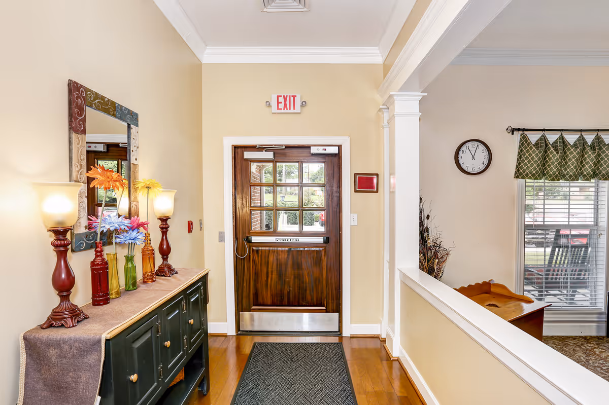 Interior entryway showing a wooden exit door with glass panes, a console table with lamps and colorful vases under a mirror, and a clock by a window.