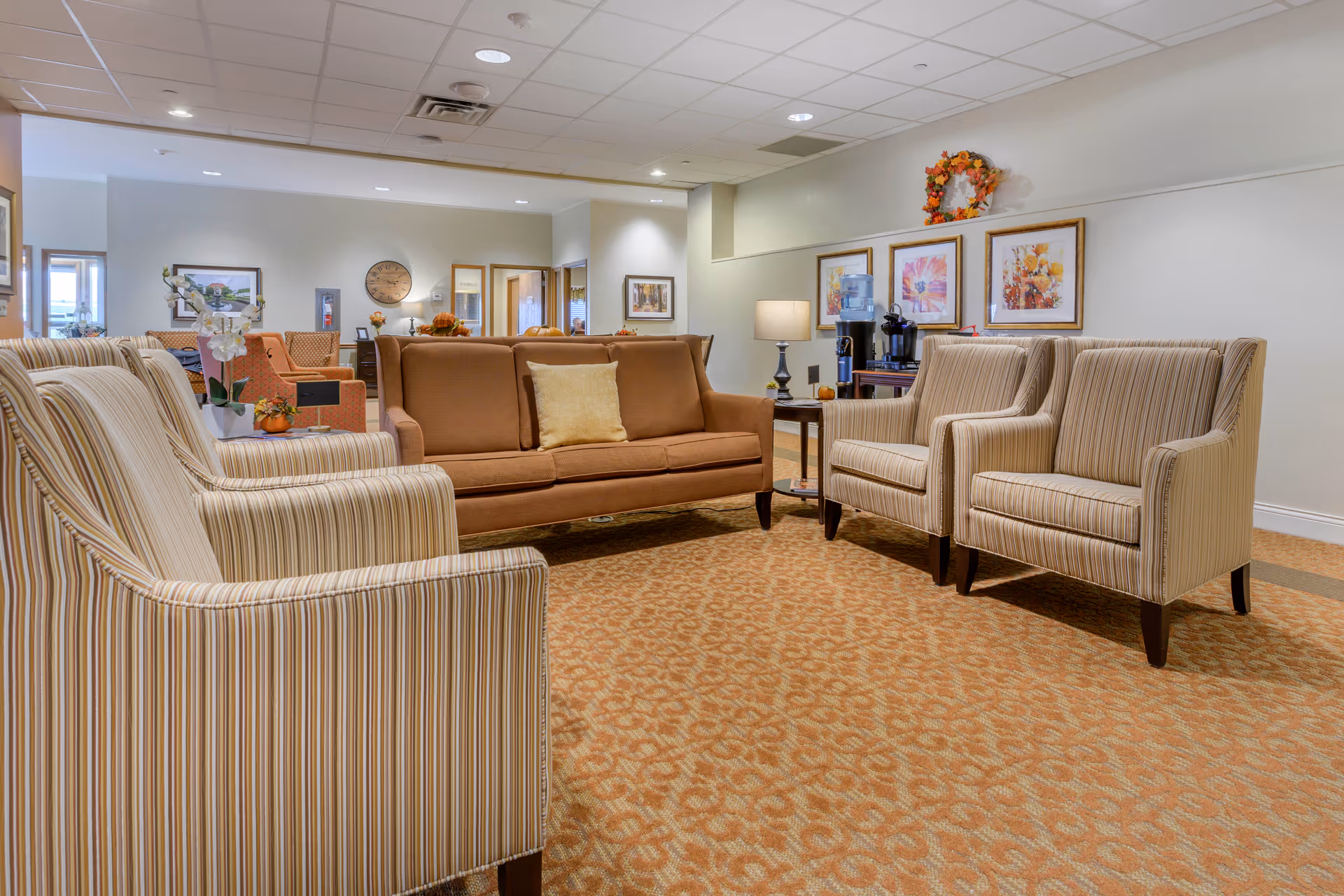 A cozy senior living facility lounge area with a brown sofa, four striped armchairs, a side table with a lamp, and a coffee station with a water dispenser and coffee maker. The walls are decorated with framed floral artwork and a fall-themed wreath. The carpet has a patterned design and the ceiling has recessed lighting.