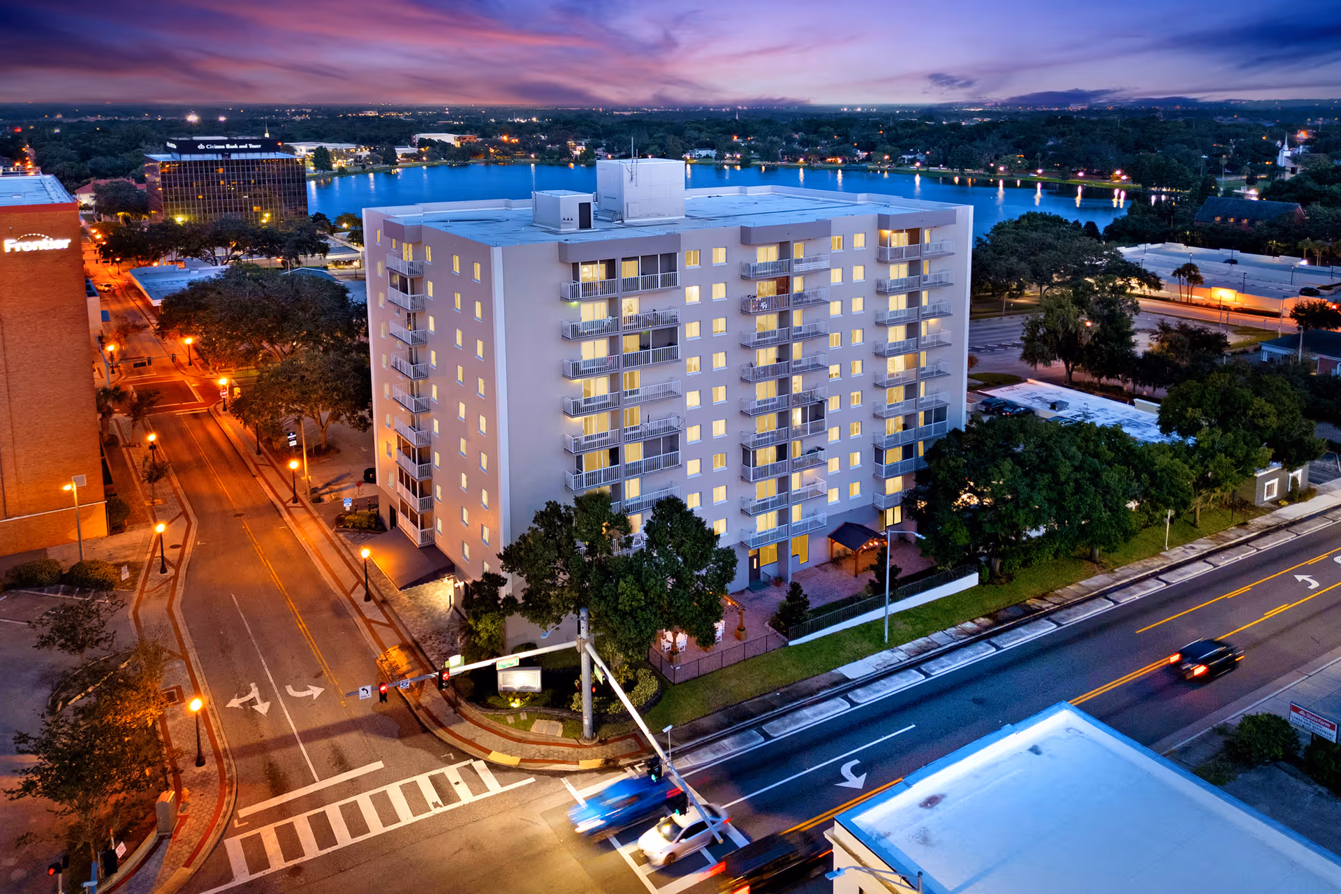 Aerial view of a multi-story residential building named Lake Morton Plaza at dusk, with illuminated windows and balconies. The building is situated near a lake with city lights and trees surrounding the area. Streets with traffic lights and cars are visible in the foreground.