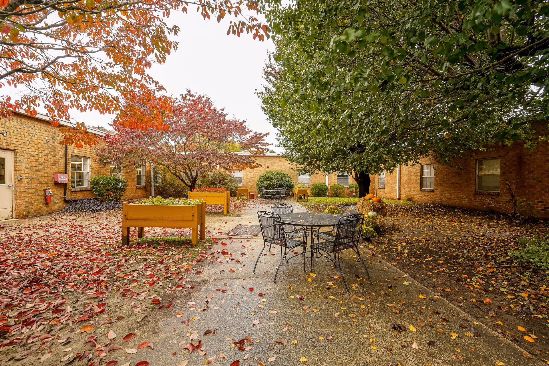 Courtyard patio surrounded by brick buildings with a metal table and chairs, raised planters, and autumn leaves on the ground.
