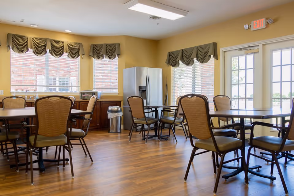 A bright dining room with several tables and chairs arranged on a wooden floor. The room has large windows with green valances, a stainless steel refrigerator, a trash can, and a microwave on a countertop. There is an exit door with glass panels letting in natural light.