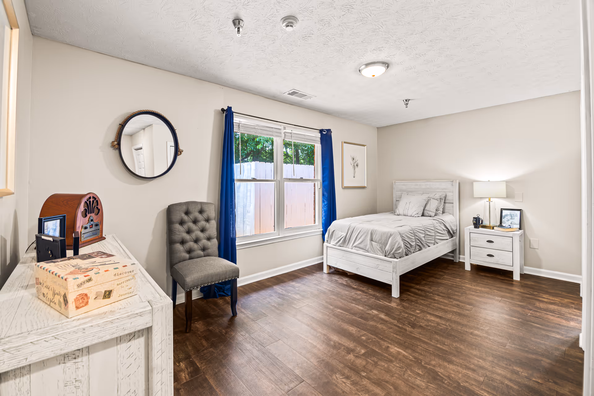 A bright and clean bedroom with a single bed dressed in light gray bedding, a matching nightstand with a lamp, a framed picture, and a mug. There is a gray upholstered chair next to a window with blue curtains, and a white wooden dresser with decorative items including a vintage-style radio and a decorative box. The room has wood flooring and light-colored walls.