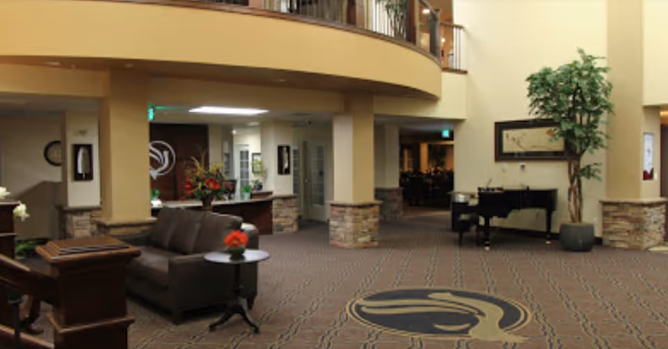 Spacious lobby area with a brown leather sofa and a small round table with a red flower vase. The floor is carpeted with a patterned design featuring a large logo in the center. There is a grand piano near a large potted plant and framed artwork on the wall. The lobby has a high ceiling with a balcony railing above and stone accents on the columns.