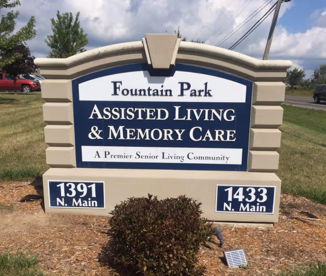 Outdoor sign for Fountain Park Assisted Living & Memory Care, a premier senior living community, with addresses 1391 N. Main and 1433 N. Main visible on the sign. The sign is set on a grassy area with a small bush in front and a partly cloudy sky in the background.