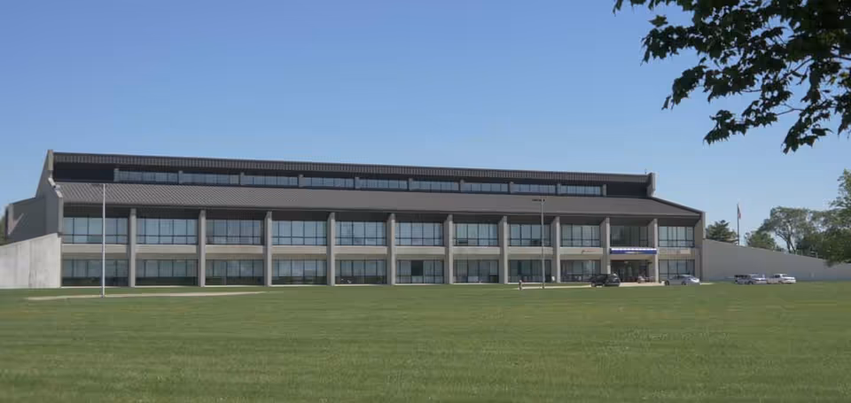 A large, modern three-story building with many windows and a sloped roof, situated behind a wide expanse of green grass under a clear blue sky. A few cars are parked near the entrance on the right side, and a tree branch is visible in the upper right corner.