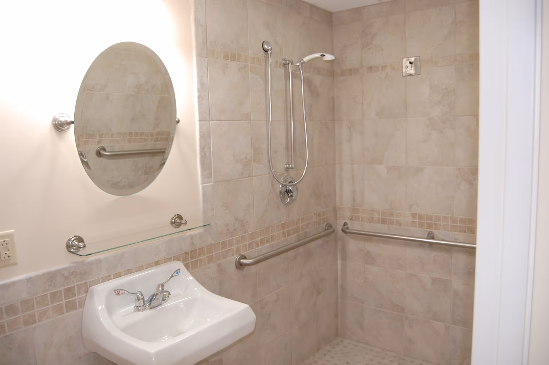 A bathroom with beige tiled walls and floor, featuring a wall-mounted sink with hot and cold faucets, a round mirror above the sink, a glass shelf, and a walk-in shower area with a handheld showerhead and multiple grab bars for accessibility.