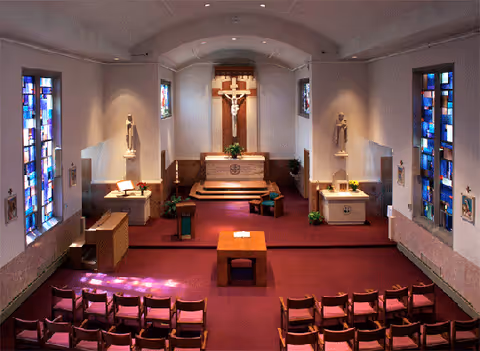 Interior view of a chapel with rows of chairs, stained glass windows, an altar, and a crucifix.