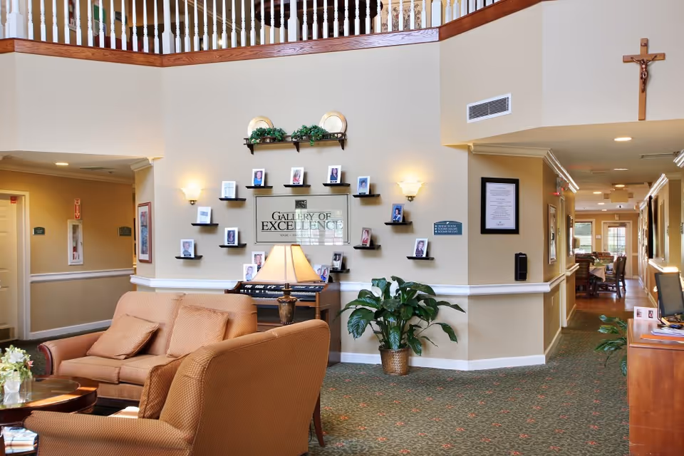 Seated lounge area with armchairs and a lamp facing a wall of small photo shelves and a 'Gallery of Excellence' sign in a senior living facility lobby.