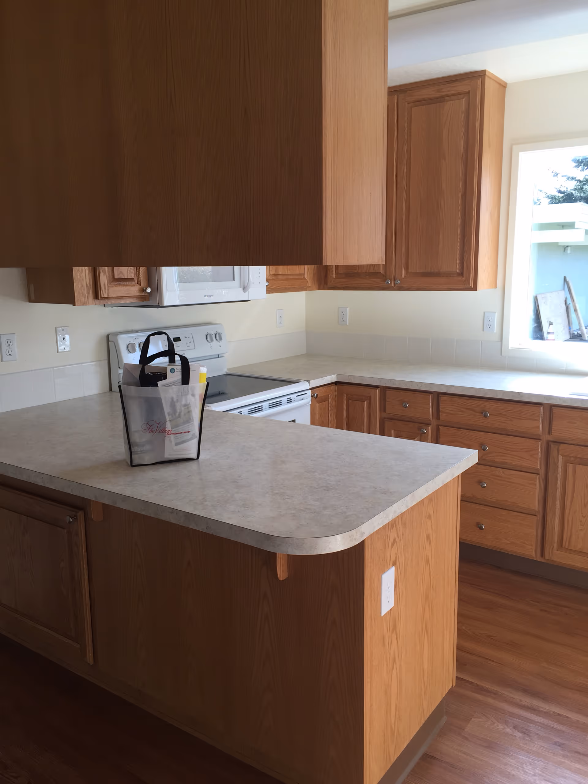Sunlit kitchen with wood cabinets, a countertop island, stove and microwave, and a tote bag on the counter.