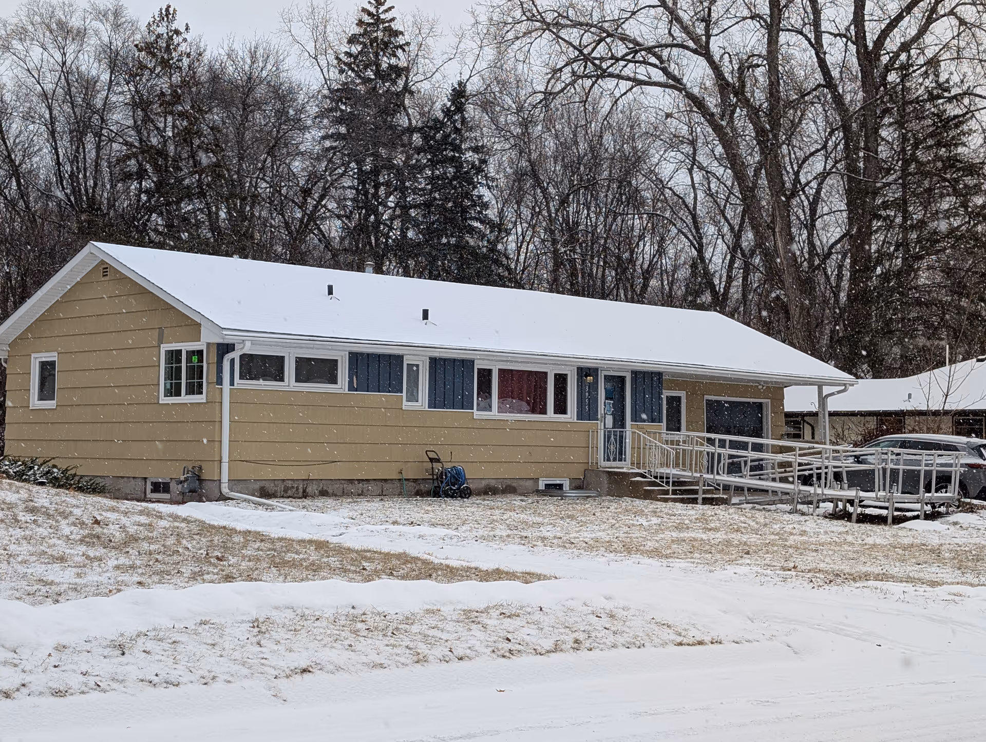 Single-story yellow ranch house with a snow-covered roof, wheelchair ramp, and trees in the background during snowfall.