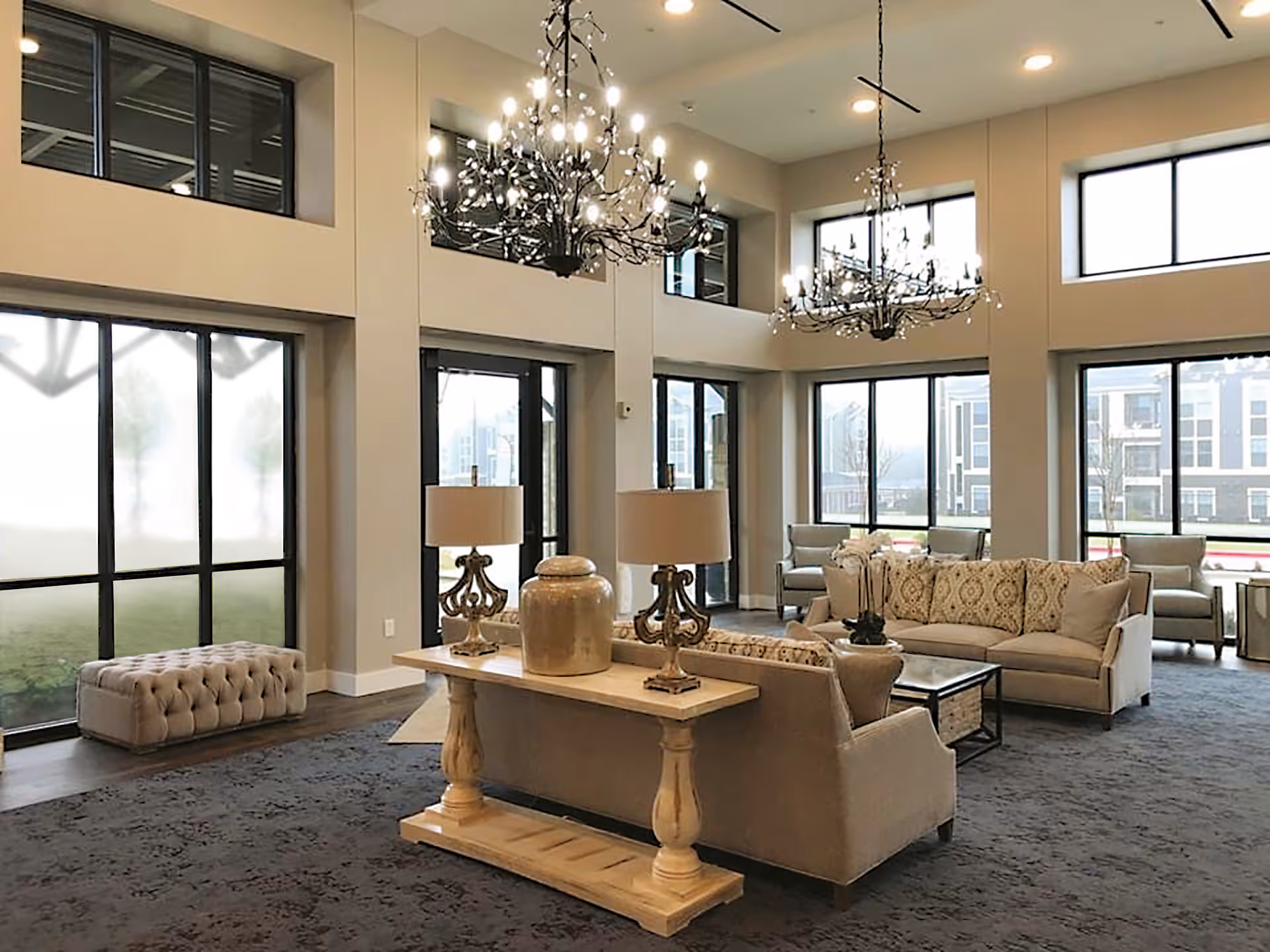 A spacious and well-lit living room area with large windows, two chandeliers, beige sofas with patterned cushions, a wooden console table with two lamps and a decorative vase, and a tufted bench near the window.