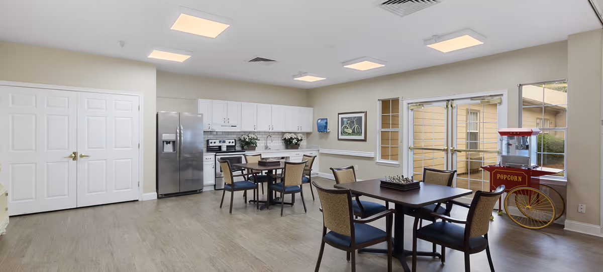 A bright and clean communal kitchen and dining area with white cabinets, stainless steel refrigerator, stove, and two tables with chairs. A popcorn machine is placed near glass doors leading outside. The room has light-colored walls and flooring, with ceiling lights providing illumination.