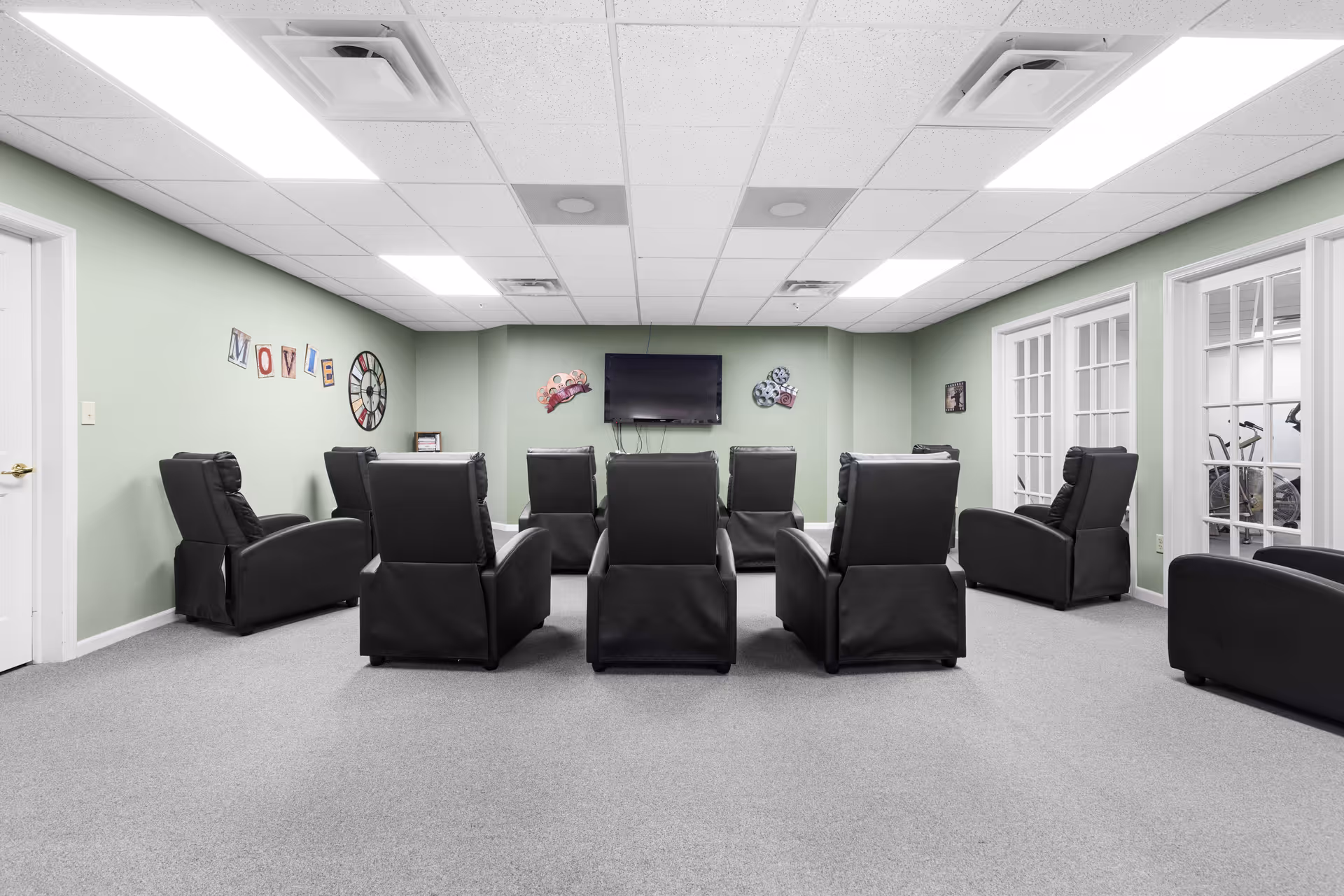 A room with eight black recliner chairs arranged in two rows facing a wall-mounted flat screen TV. The walls are painted light green and decorated with movie-themed wall art, including the word 'MOVIE' and film reels. The room has a gray carpet and a white drop ceiling with fluorescent lights. There are glass-paneled doors on the right side leading to another room with exercise equipment.