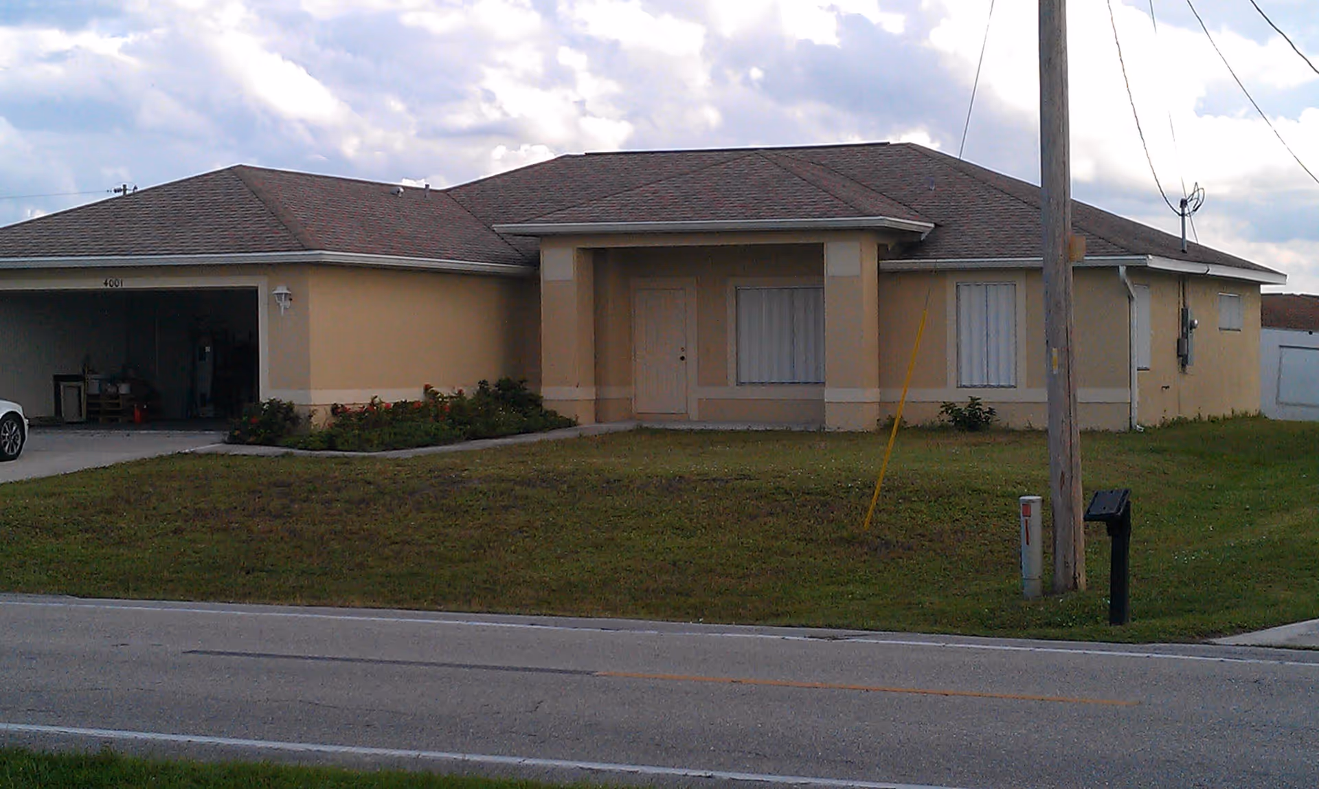 Single-story beige residential building with a brown shingled roof, a front door with a small covered porch, two windows with closed blinds, an attached garage with an open door, a grassy front yard, a mailbox, and a utility pole near the street.