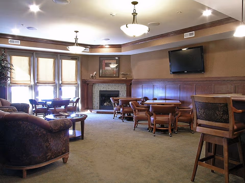 A cozy common area with multiple seating options including armchairs, small tables, and chairs arranged around a fireplace. The room features warm brown tones, a wall-mounted flat screen TV, and large windows with shades allowing natural light to enter.