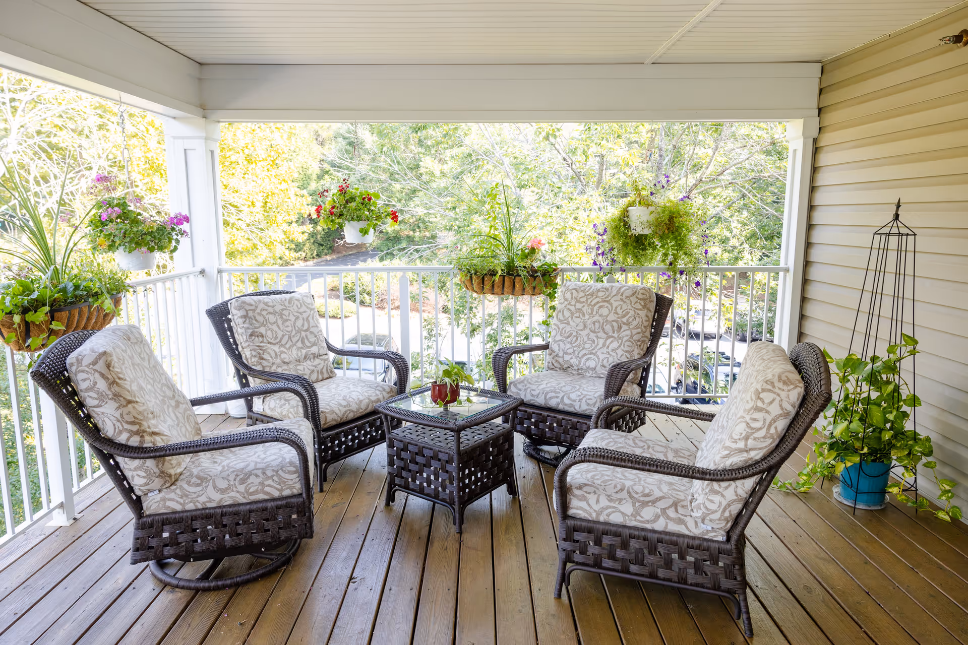 Covered outdoor patio area with four cushioned wicker chairs arranged around a small glass-top wicker table. The patio has wooden flooring and white railings with hanging flower pots and potted plants placed around the space. Trees and greenery are visible in the background.
