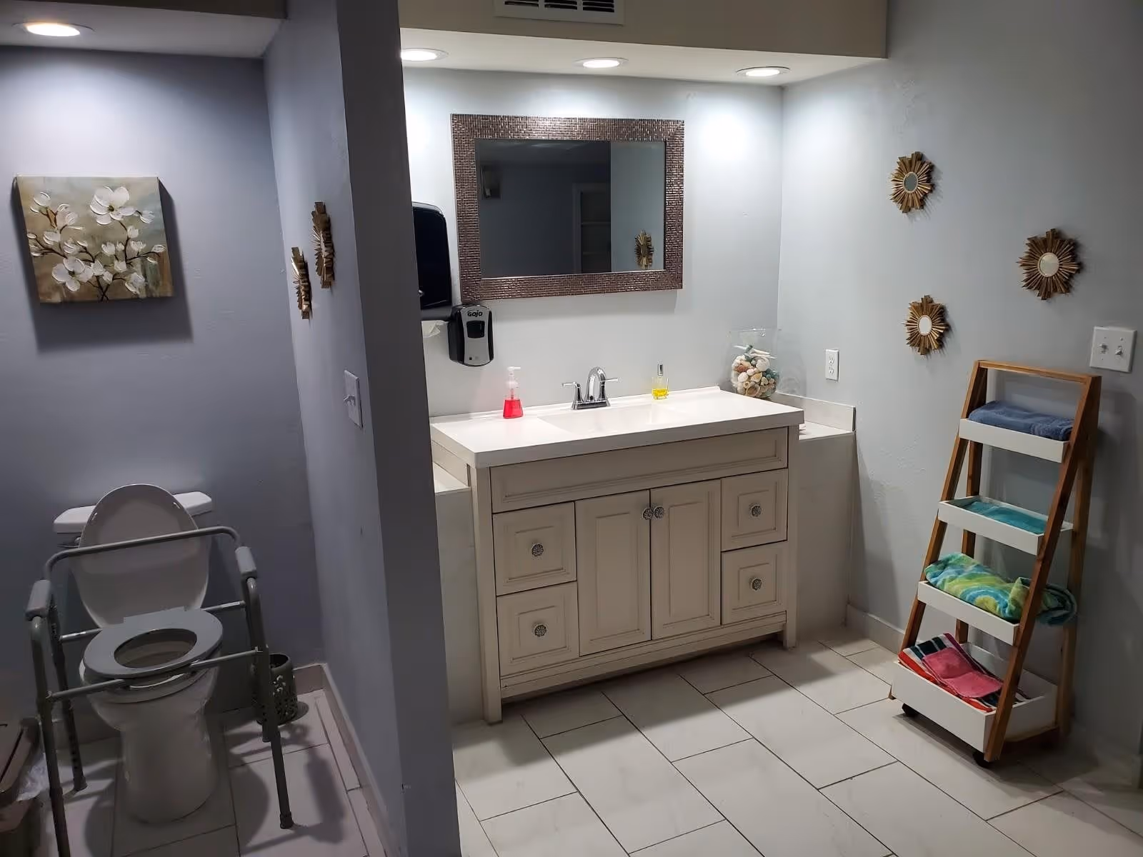 A clean and well-lit bathroom with a white toilet equipped with safety rails on the left side, a white vanity with a sink and a large mirror above it in the center, and a wooden towel rack holding colorful towels on the right side. The walls are painted light gray and decorated with floral artwork and small sunburst mirrors.