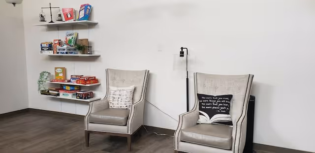 A quiet sitting area with two beige armchairs, each with a decorative pillow. One pillow has a quote, and the other has a musical notes pattern. To the left, there are three wall-mounted shelves holding board games, books, and small decorative items. The floor is wooden, and the walls are plain white.
