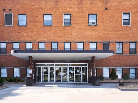 Front exterior view of a brick building with multiple windows and a covered entrance with glass doors. There are trash bins and small shrubs near the entrance.