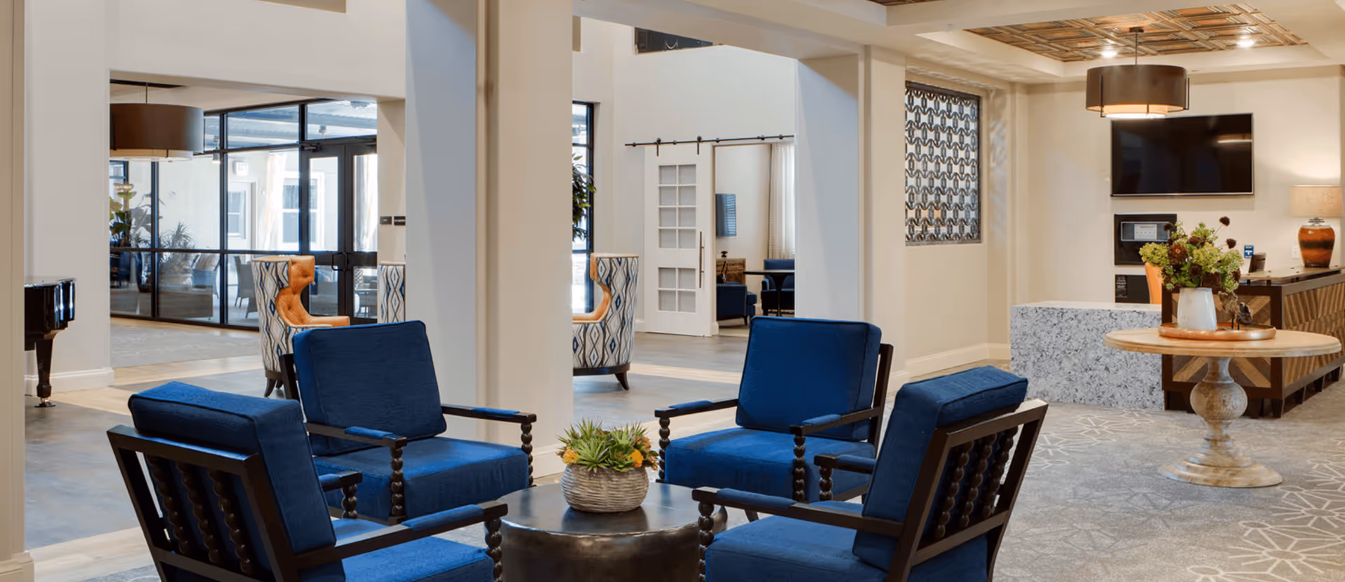 A spacious and well-lit common area with four blue cushioned armchairs arranged around a small round table with a plant centerpiece. The room features light-colored walls, a patterned carpet, a round wooden table with a flower arrangement, a flat-screen TV mounted on the wall, and a piano in the background near large glass doors.