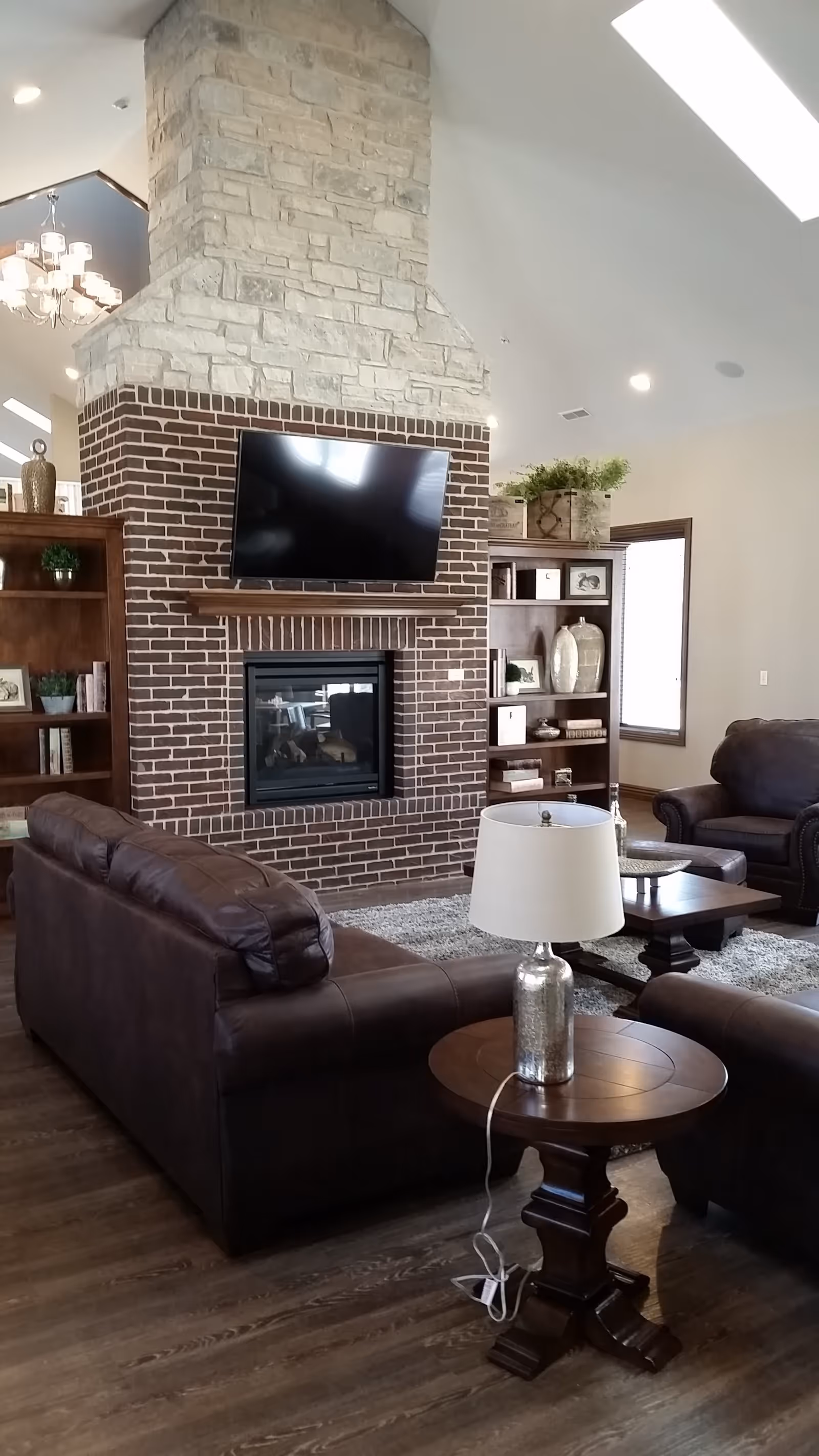 Spacious living room with leather sofas, a brick fireplace topped by a mounted TV, bookshelves, and a side table with a lamp.