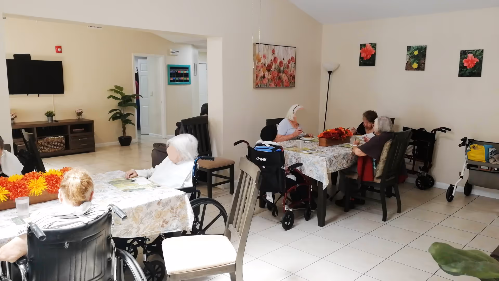 A group of elderly individuals sitting around two tables in a well-lit room with tiled floors and light-colored walls. Some are in wheelchairs, and the tables are covered with floral tablecloths and decorated with orange and yellow flowers. The room has a TV mounted on the wall, a plant, and several floral paintings on the walls. Mobility aids such as walkers are visible in the background.