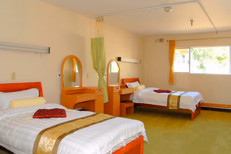 A bright room with two single beds, each with white bedding, a gold and brown decorative runner, and a red cushion. Between the beds are two wooden dressers with mirrors and small stools. A green curtain hangs from the ceiling between the beds, and a large window with an orange curtain lets in natural light.