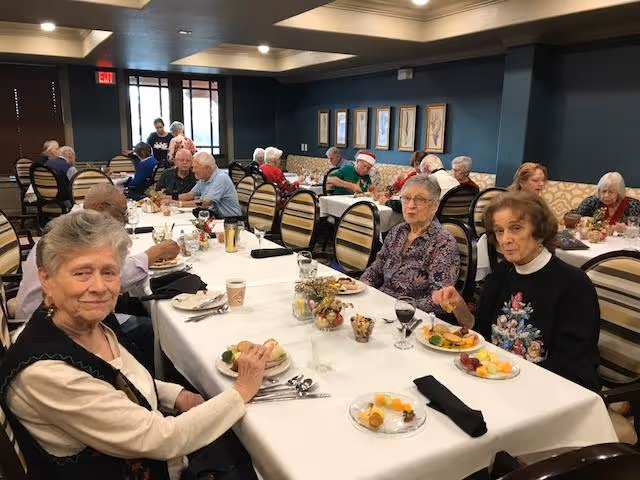 A group of elderly people seated at long dining tables in a well-lit room, enjoying a meal together. The tables are covered with white tablecloths and set with plates of food, glasses, and utensils. The room has dark walls with framed artwork and a patterned cushioned bench along one side.