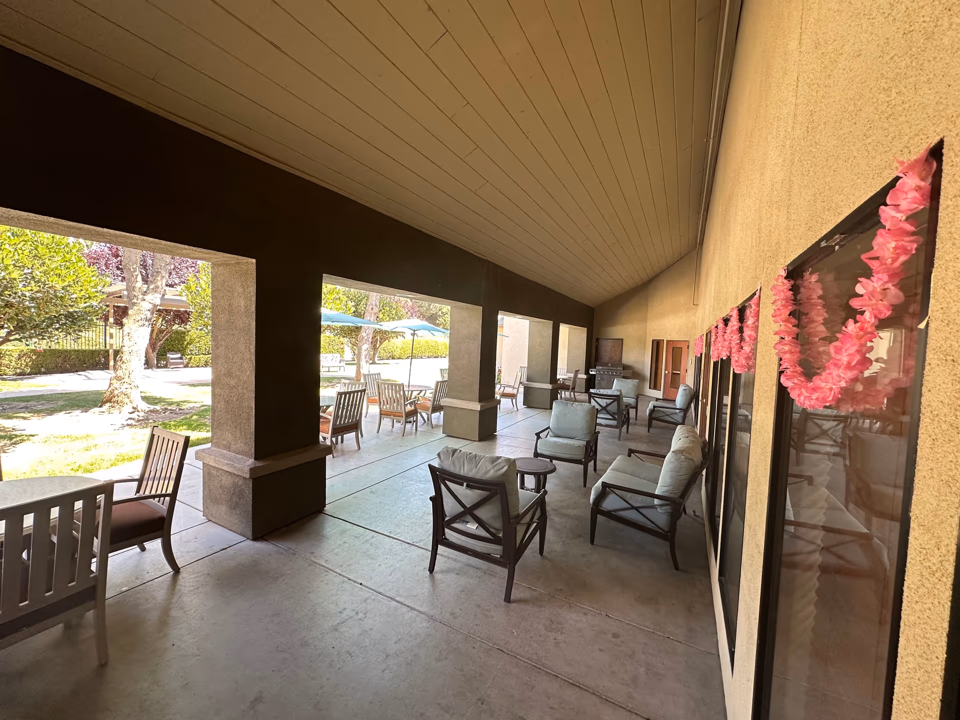 Covered outdoor patio area with cushioned chairs and small tables, decorated with pink garlands on the wall. Beyond the patio, there are tables with umbrellas and chairs on a grassy area with trees and bushes.