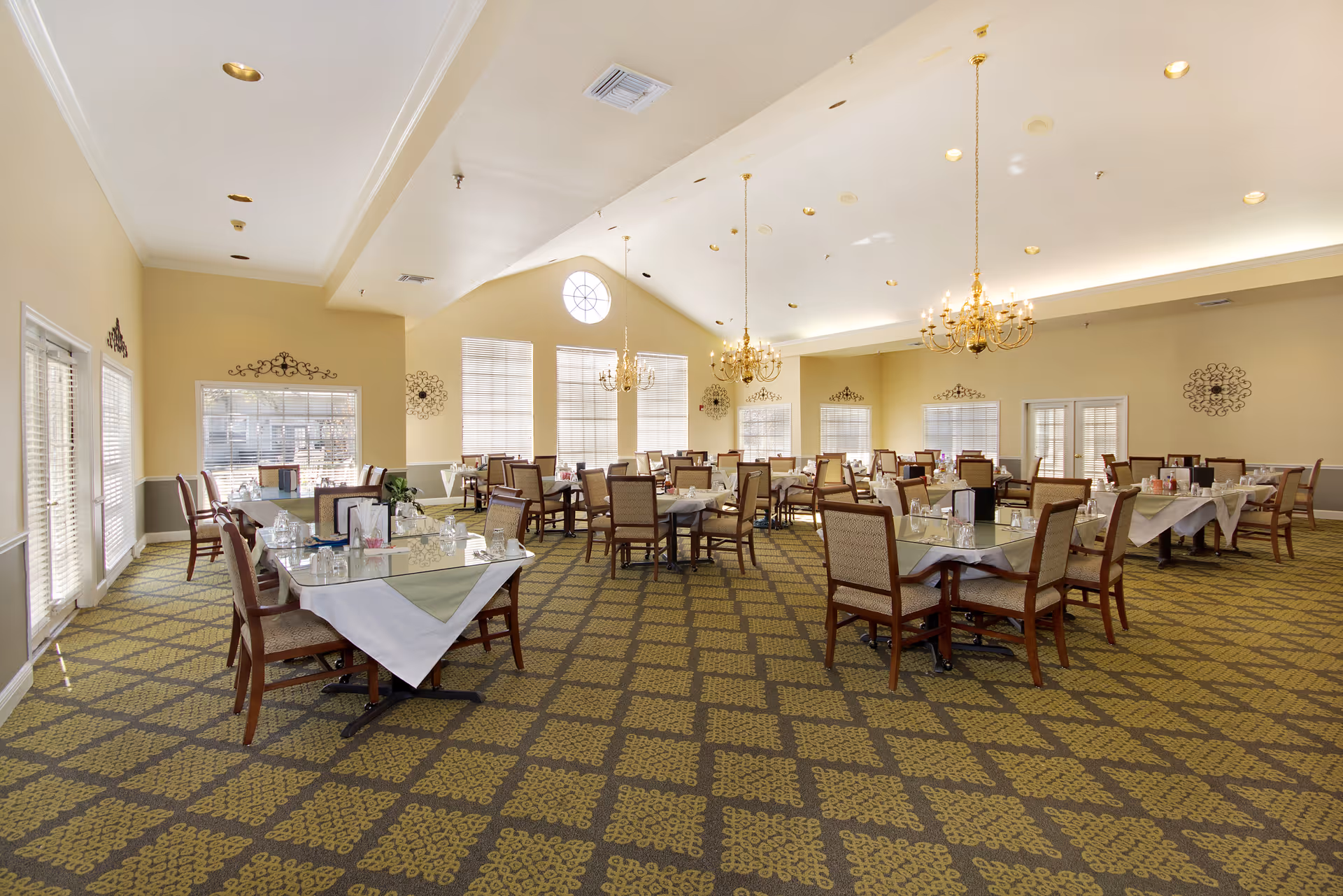 A spacious dining room with multiple tables covered with white tablecloths and set with glassware and napkins. The room features large windows allowing natural light, chandeliers hanging from a high ceiling, and patterned carpet flooring.